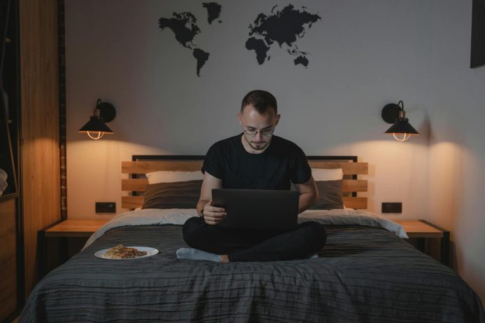 Man working on a laptop in a cozy bedroom setting, showcasing remote work lifestyle.